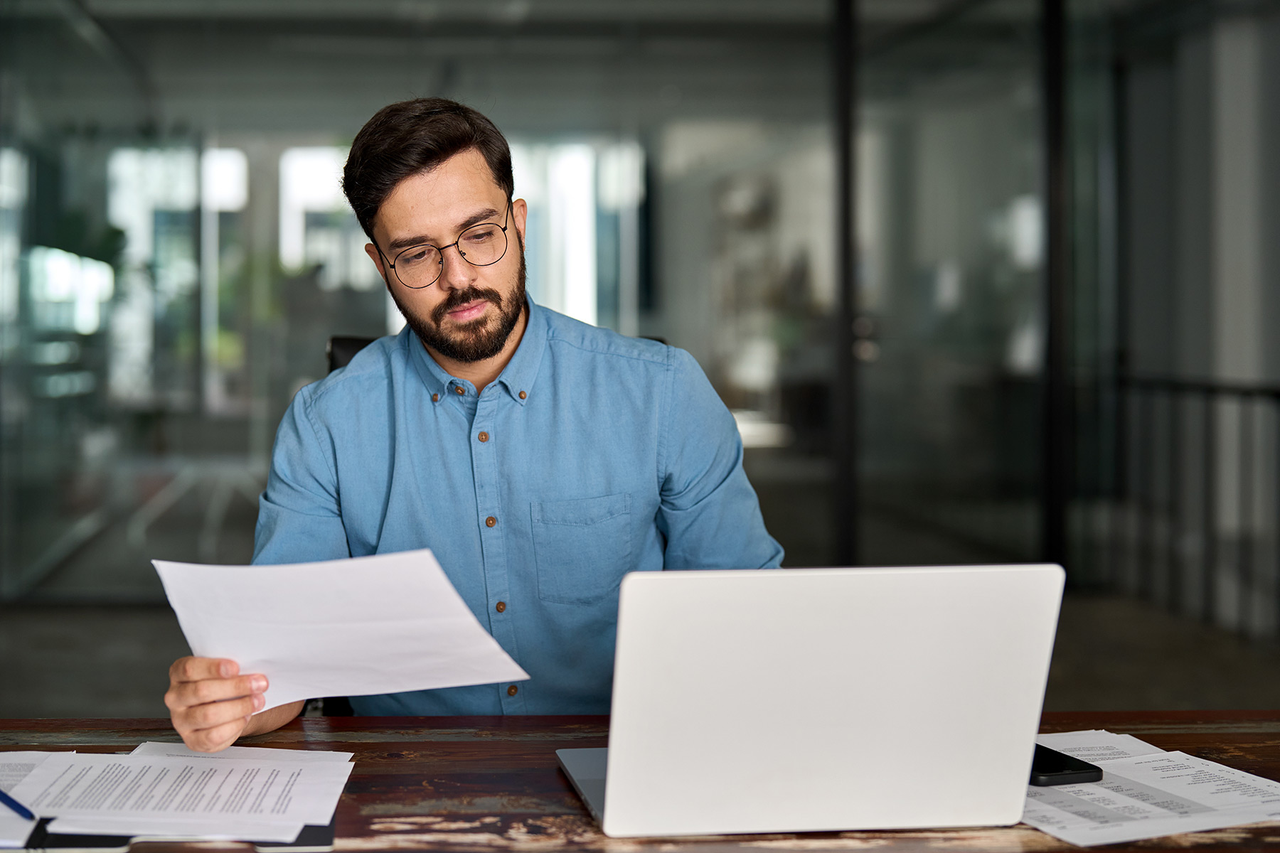 A person sits at a desk in an office, holding and looking at a piece of paper. An open laptop and several documents are on the desk. The background includes glass walls and office furniture.