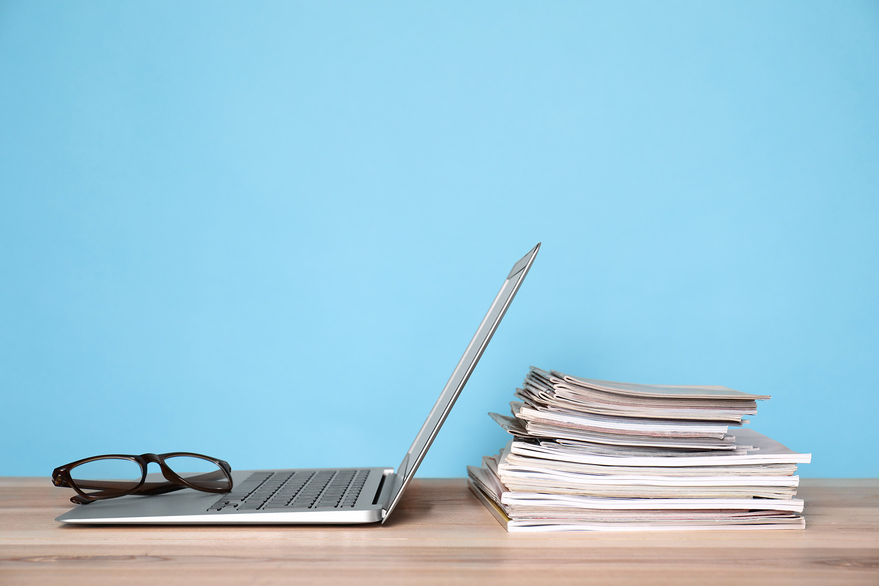 A partially open laptop with a pair of eyeglasses resting on the keyboard sits on a desk next to a stack of papers or magazines, against a plain background.