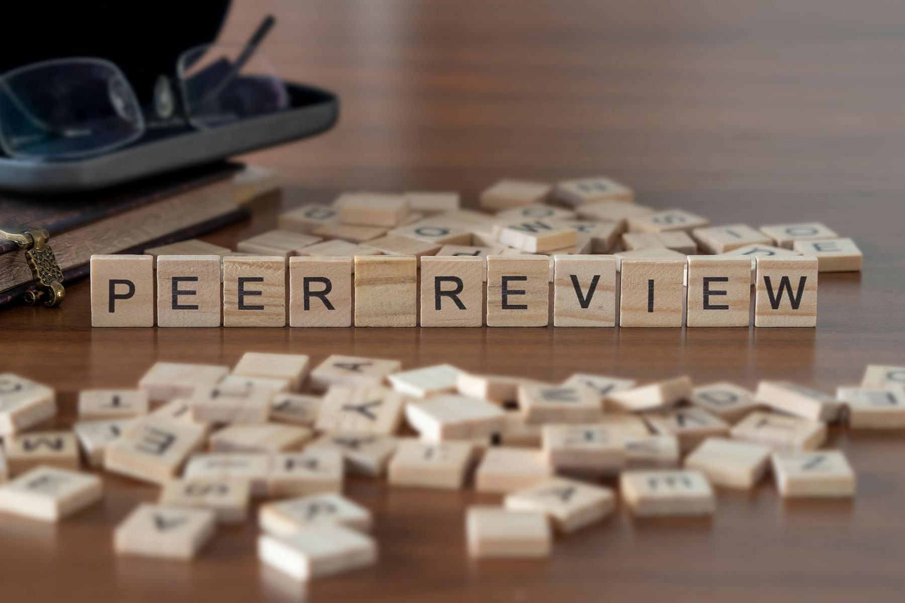 Wooden letter tiles spelling 'PEER REVIEW' on a surface, with scattered tiles and a pair of glasses in the background.