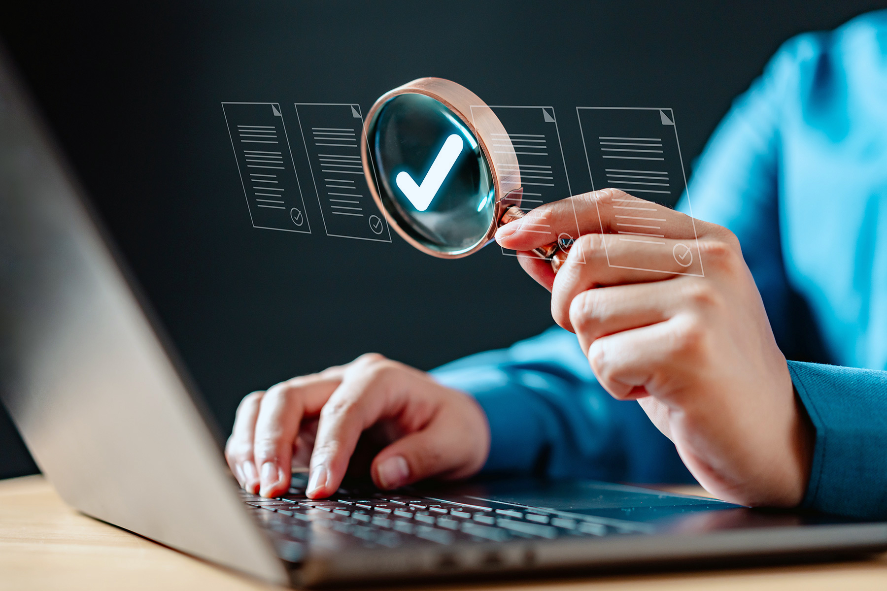 A person uses a magnifying glass to examine digital documents with check marks, while typing on a laptop.